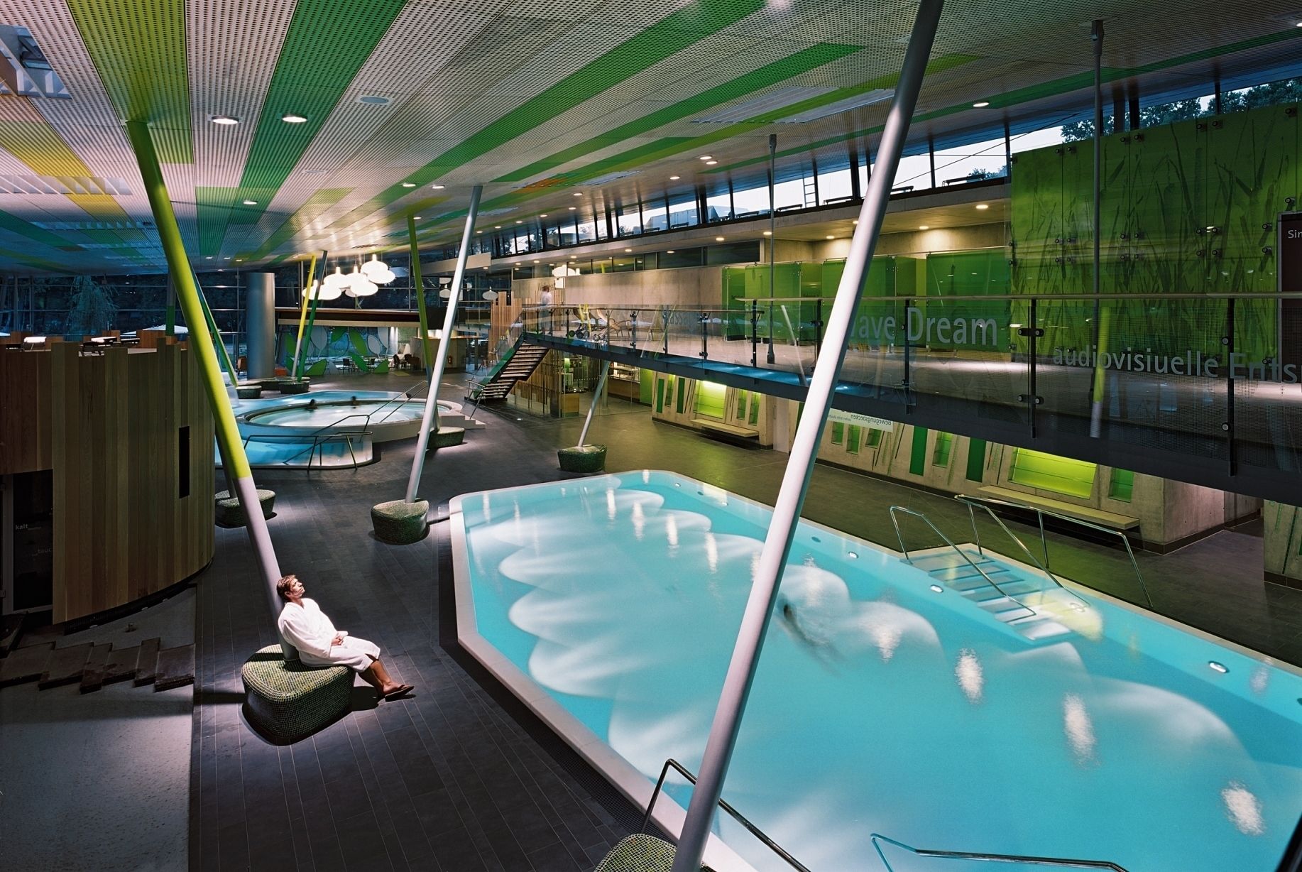 A modern indoor swimming pool with multiple levels, a central seating area, and a green wall with text.