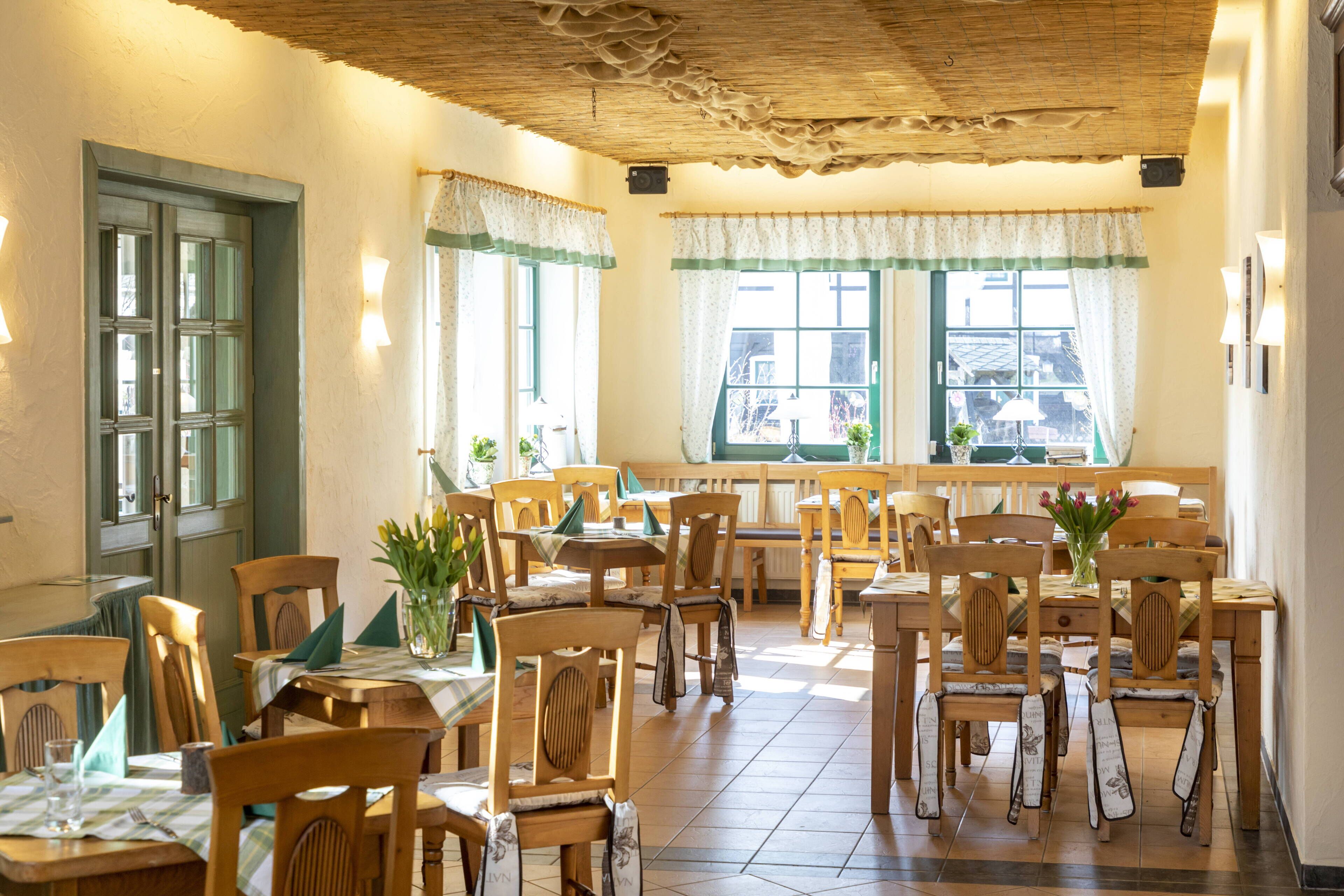 A dining area with wooden tables and chairs, large windows with curtains, and a rustic ceiling.
