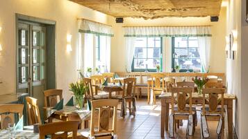 A dining area with wooden tables and chairs, large windows with curtains, and a rustic ceiling.