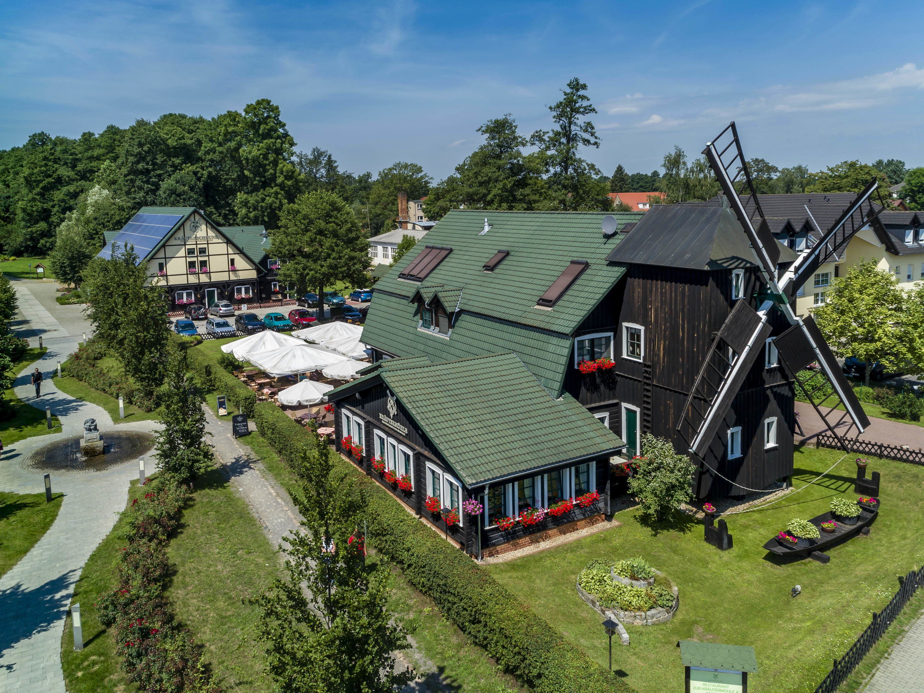 Eine traditionelle Windmühle mit einem grünen Dach, neben einem Gebäude ebenfalls mit grünem Dach und weißen Fenstern, umgeben von üppiger Vegetation und einer gepflasterten Fläche mit Außenbestuhlung.