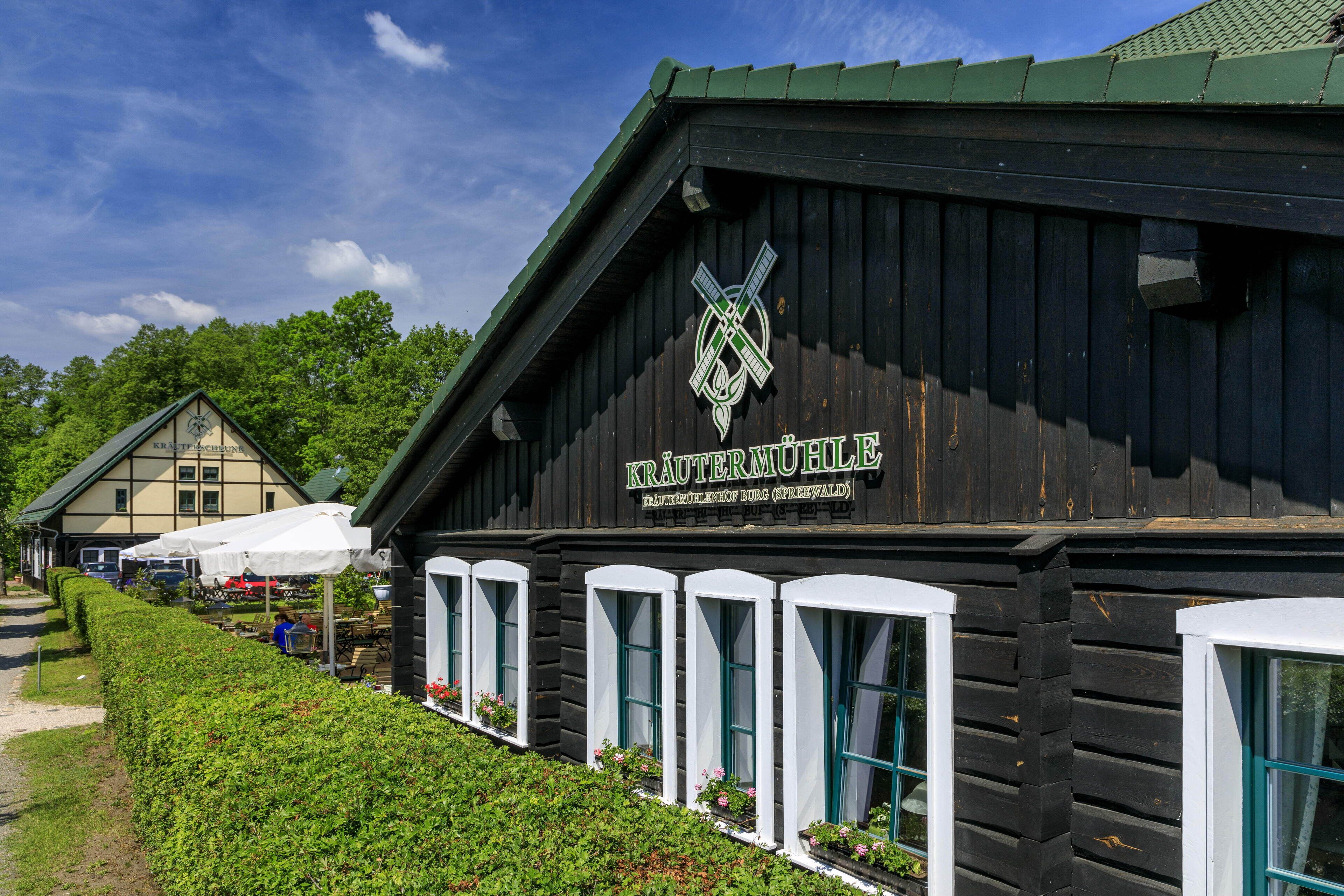 A wooden building with a sign reading "KRÄUTERMÜHLE" and a logo above the entrance. There are white awnings and a row of windows with green shutters.