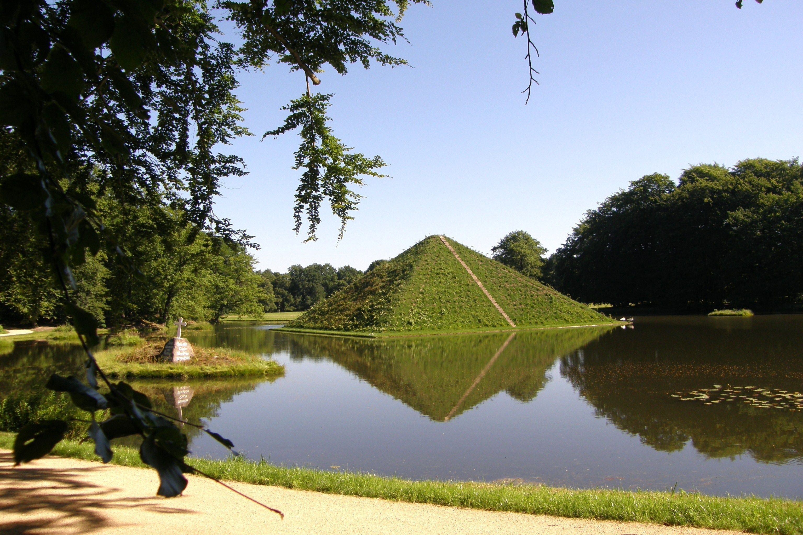 A serene landscape with a pyramid-shaped mound surrounded by a calm lake, reflecting the mound and trees.