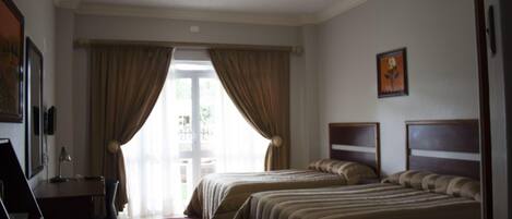 A hotel room with two beds, a desk, and a chair. There is a window with curtains, a painting on the wall, and a door leading outside.