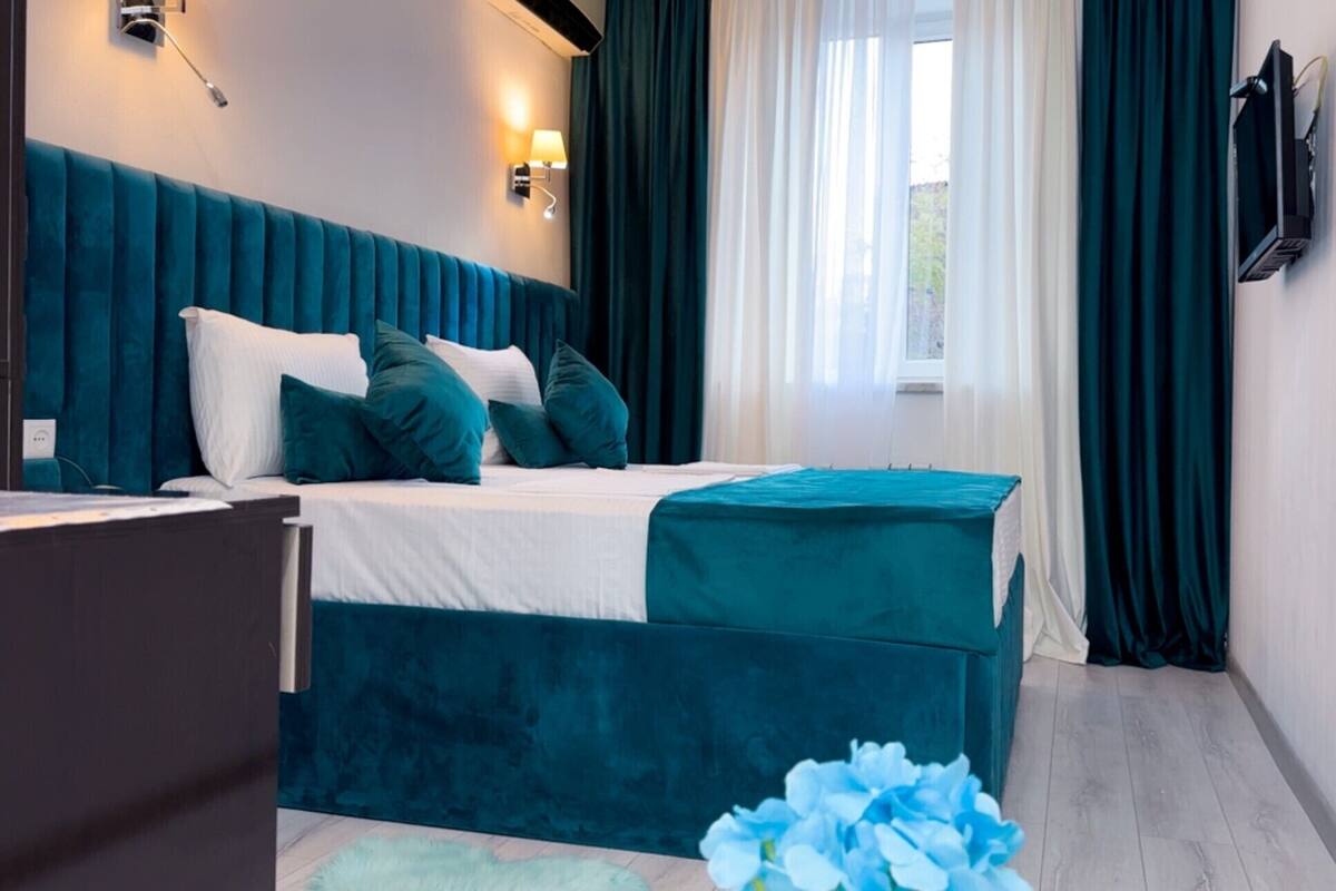Une chambre à coucher moderne avec un grand lit, une literie de couleur turquoise et une vue sur l’extérieur à travers des rideaux transparents.