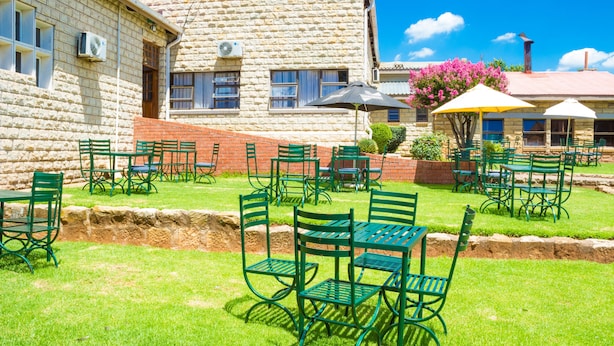 An outdoor seating area with green metal chairs and tables, a stone building with windows and air conditioning units, and a clear blue sky. - Mountain View Hotel (Hlotse)