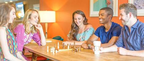 A group of people playing chess at a wooden table in a brightly lit room with orange walls and a chandelier.