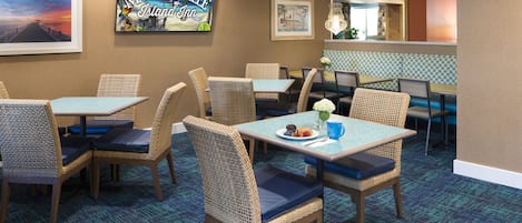 Un espace repas avec des chaises et des tables en osier, un panneau d’accueil encadré et un tapis à motifs.