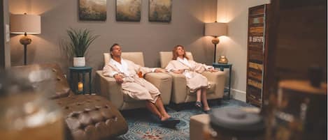 A spa room with two reclining chairs, a massage table, and a decorative wall with artwork.