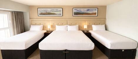 A hotel room with three single beds, a headboard with a tufted design, two framed landscape paintings, and a ceiling-mounted smoke detector.