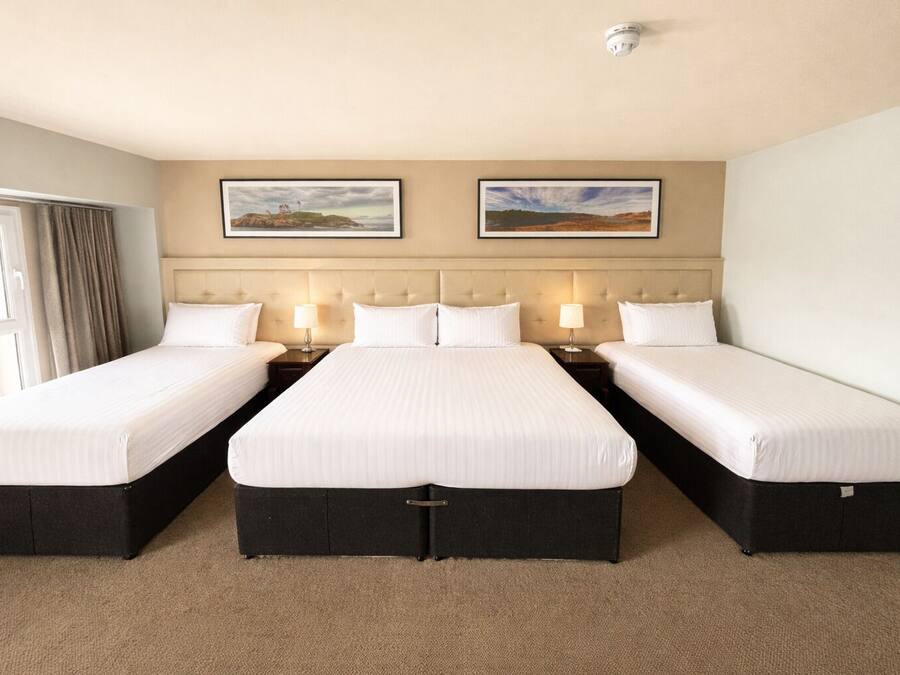 Quarto de hotel com três camas de solteiro, cabeceira com design estofado, duas pinturas emolduradas de paisagens e detector de fumaça no teto.