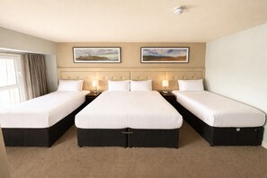 A hotel room with three single beds, a headboard with a tufted design, two framed landscape paintings, and a ceiling-mounted smoke detector.