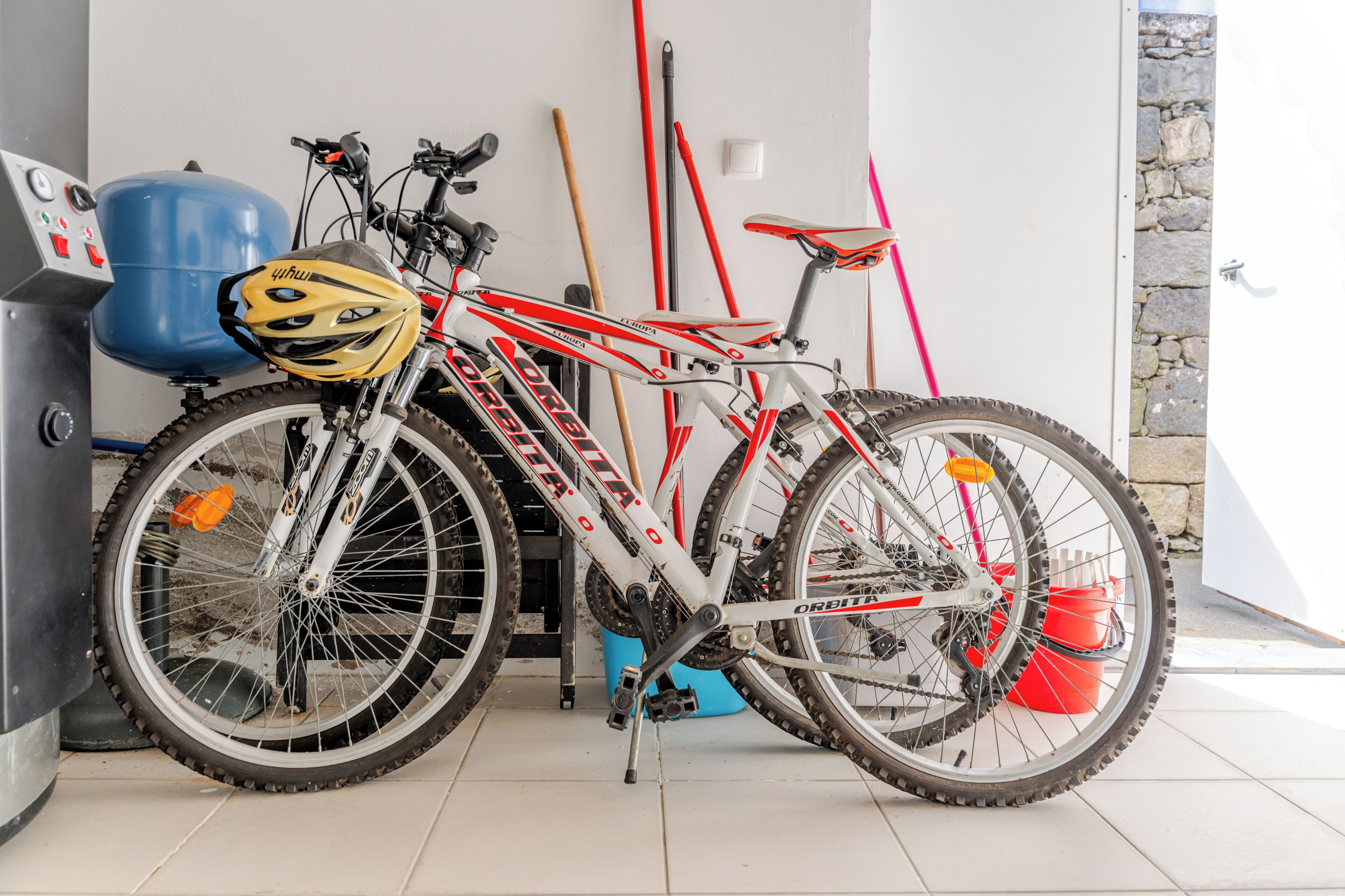 A bicycle with a helmet, a blue water heater, and a red broom leaning against a wall.