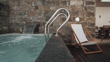 A stone wall, a metal pool ladder, a wooden chair, and a small waterfall in a pool.