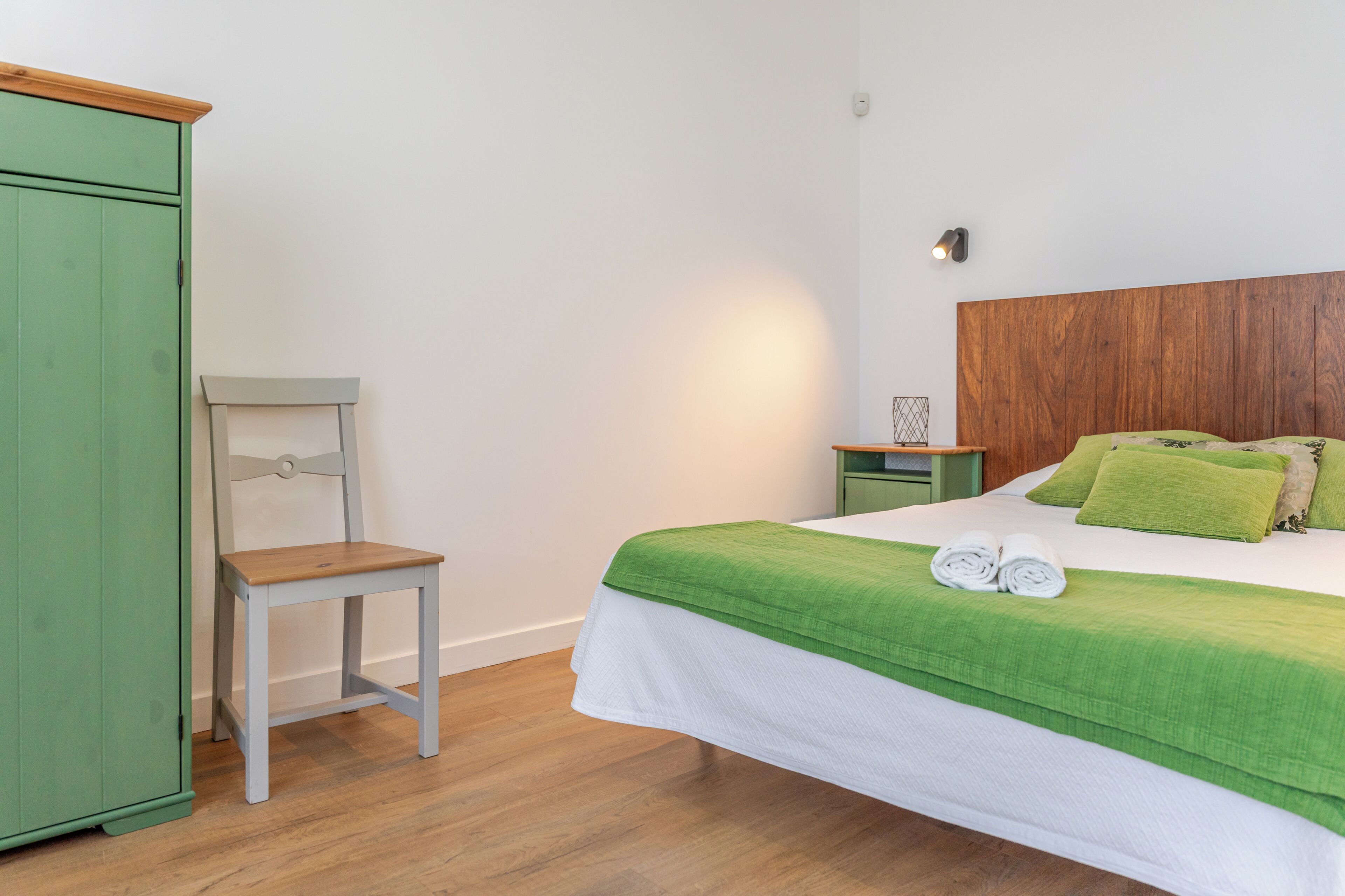 Un dormitorio con una cama, almohadas verdes, cabecera de madera, armario verde y una silla de madera.