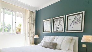 Een slaapkamer met een groot bed, een nachtkastje, een lamp en drie florale kunstwerken in lijsten aan de muur.