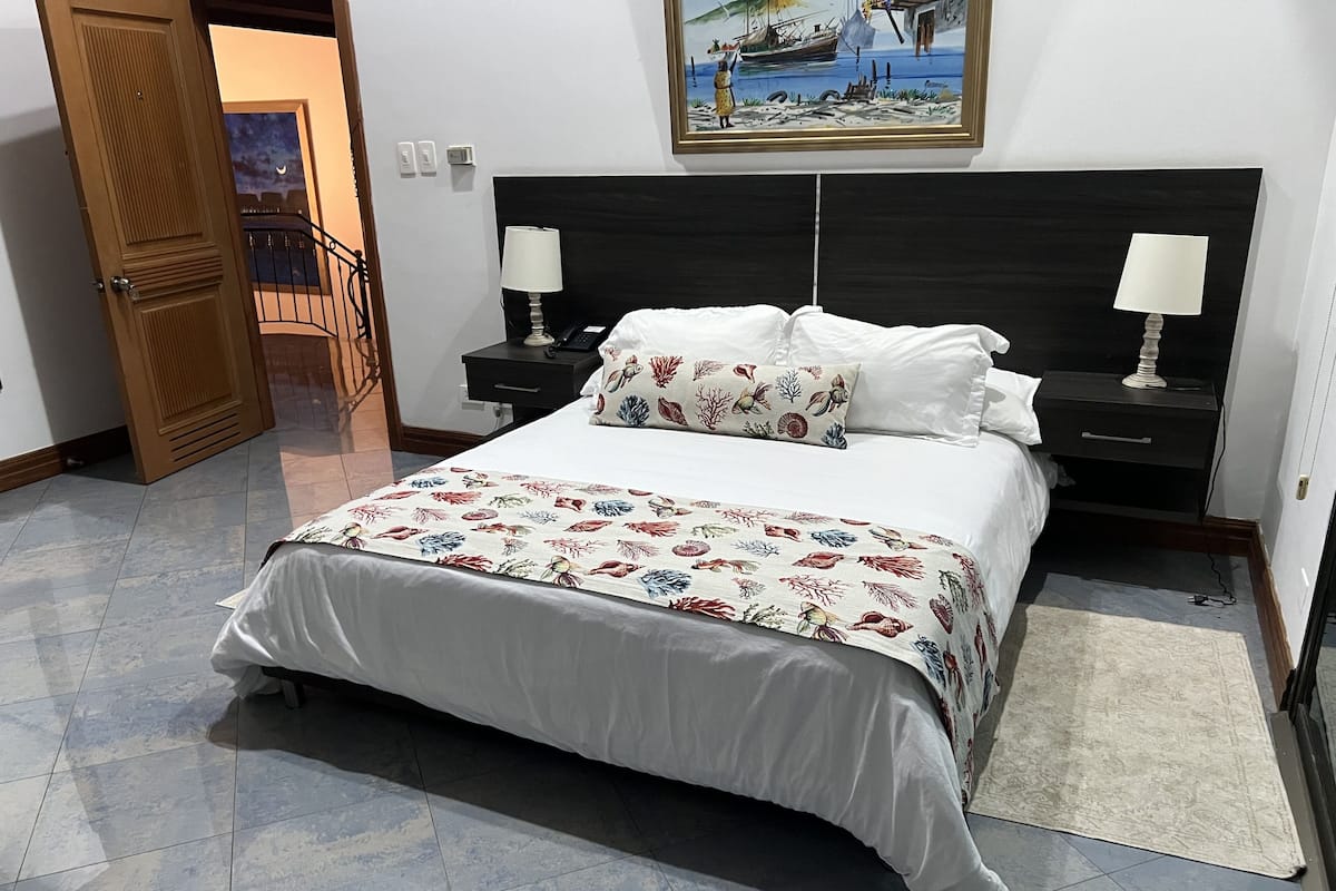 A bedroom with a large bed, two bedside tables, a nightstand, a painting on the wall, and a door leading to another room.