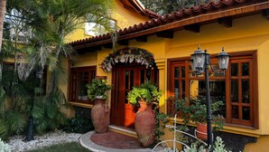 Un bâtiment jaune avec un toit en tuiles, de grandes fenêtres et un porche à l’avant, agrémenté de plantes en pot.