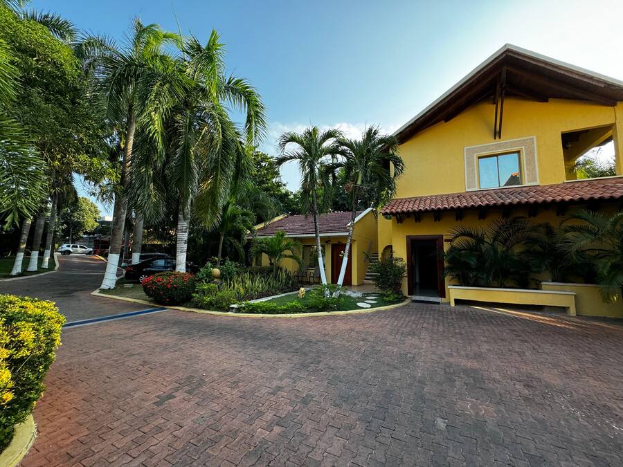 A yellow building with a tiled roof, surrounded by palm trees and a paved driveway.