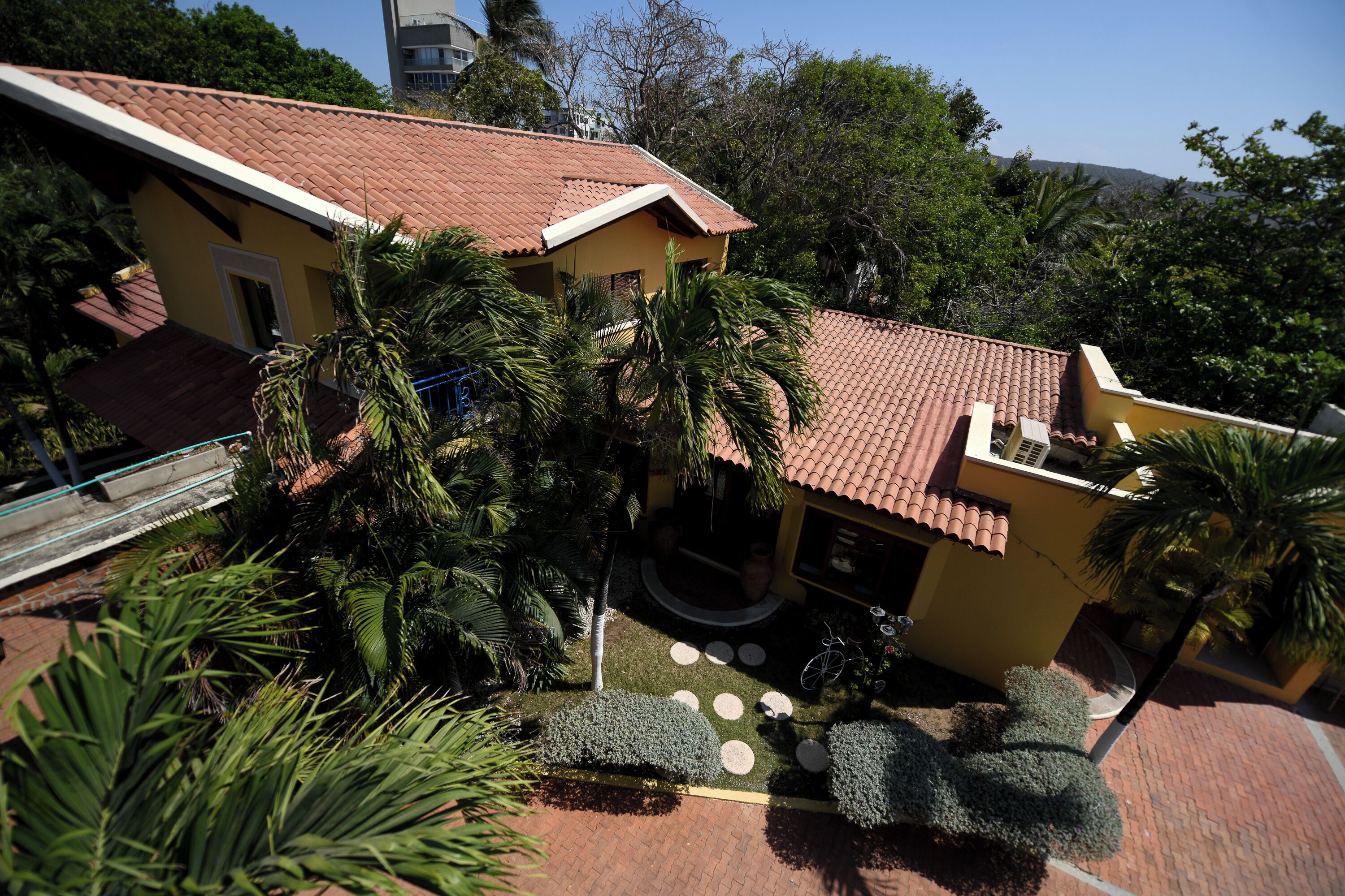 Un edificio amarillo con techo de tejas rojas, rodeado de palmeras y un patio empedrado con diseños circulares de piedra.