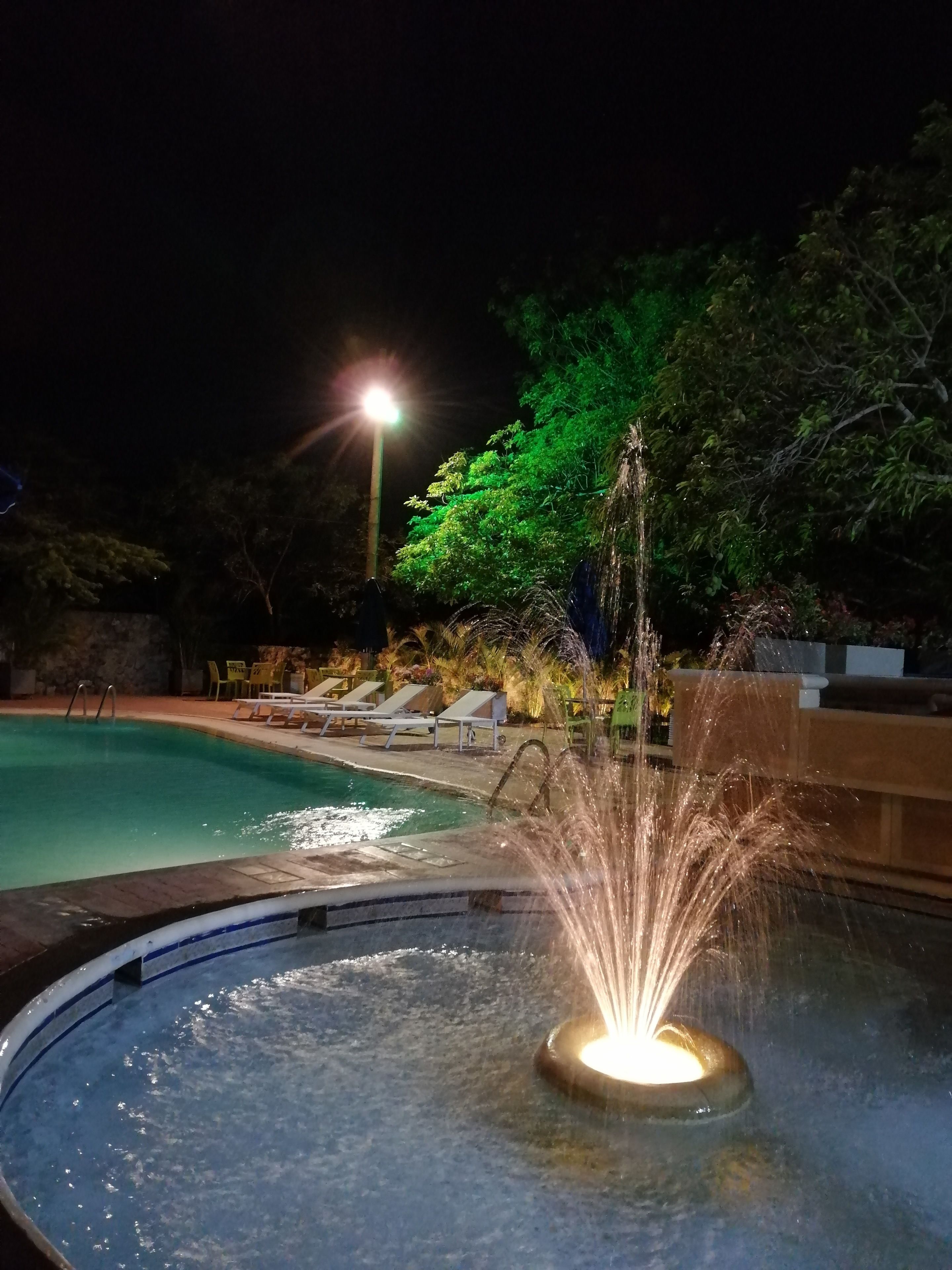 A nighttime swimming pool with a water fountain feature, surrounded by lounge chairs and a well-lit area.