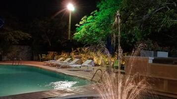 A nighttime swimming pool with a water fountain feature, surrounded by lounge chairs and a well-lit area.