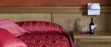 A hotel room with a wooden headboard, a bed with red patterned bedding, a nightstand with a wine bottle and glasses, and a wall-mounted lamp.