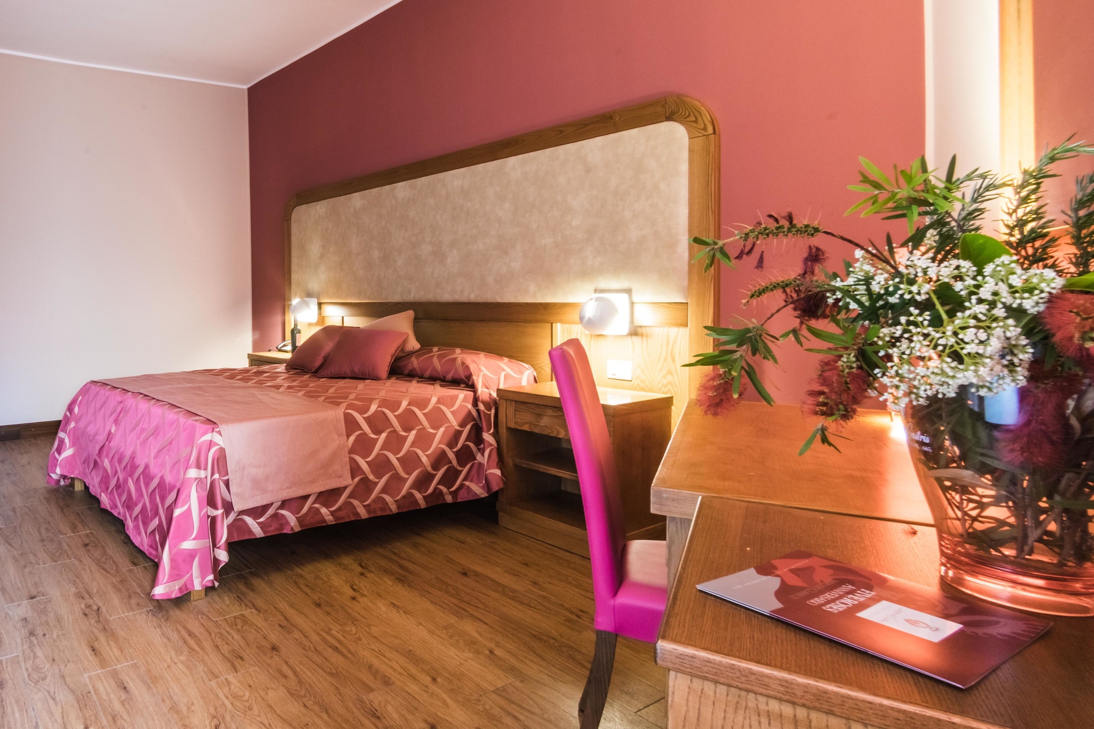 A hotel room with a bed, bedside table, chair, and a desk with a lamp and a decorative plant.