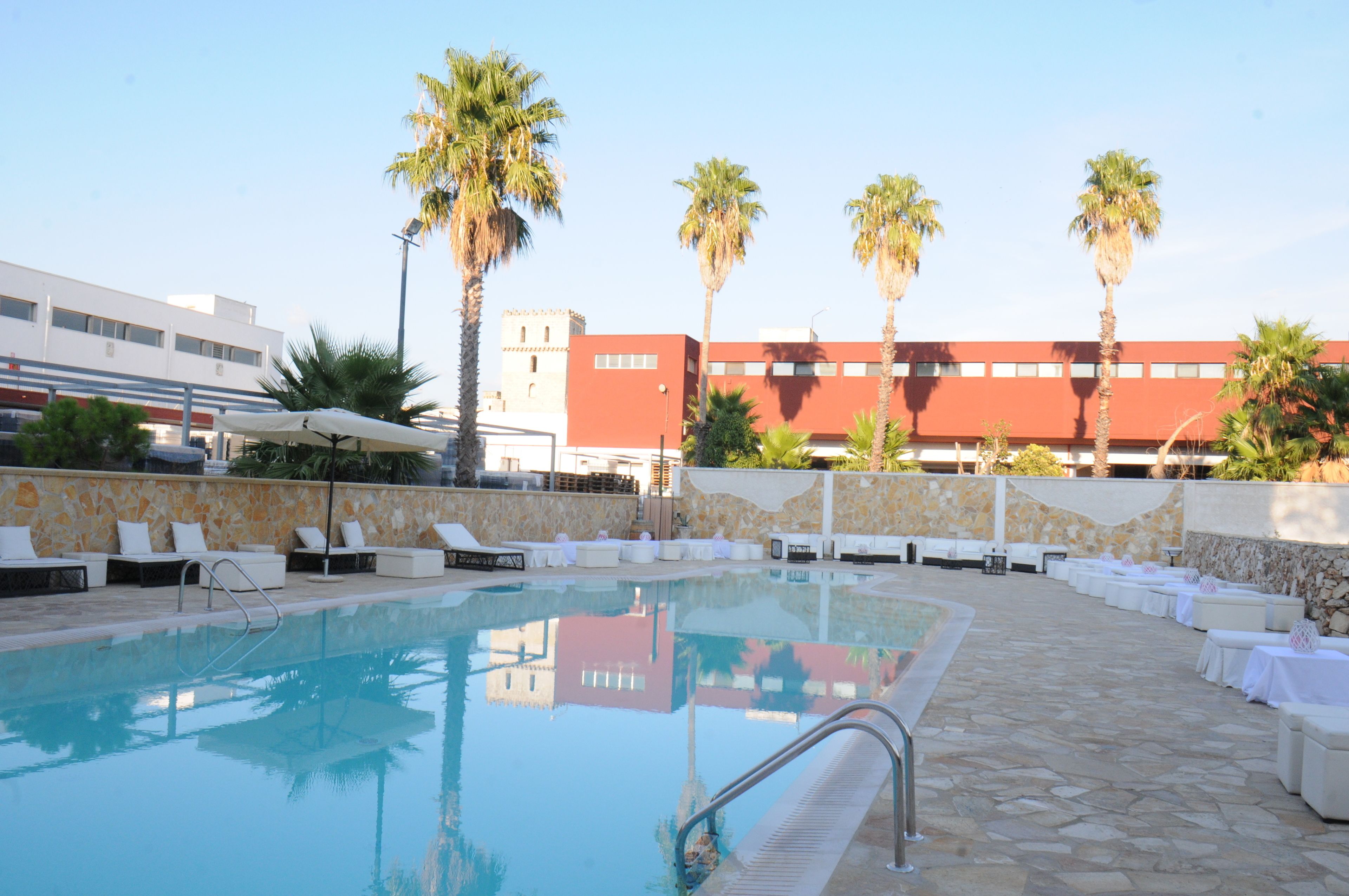 Piscina con sillas de descanso y palmeras, rodeada de un muro de piedra y un edificio de fachada roja.
