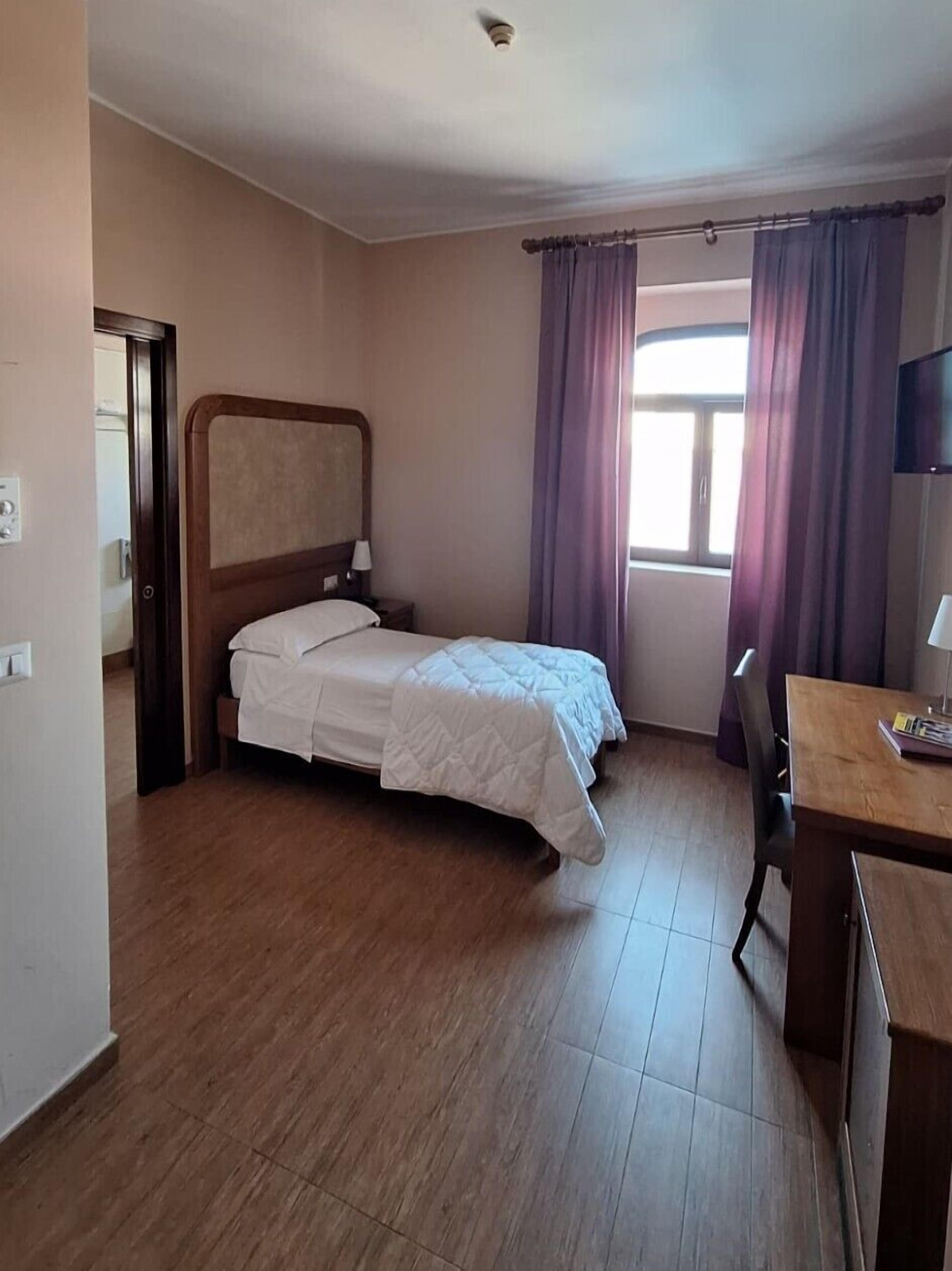 Ein Hotelzimmer mit Bett, Schreibtisch, Stuhl, Fernseher und einem Fenster mit Vorhängen.