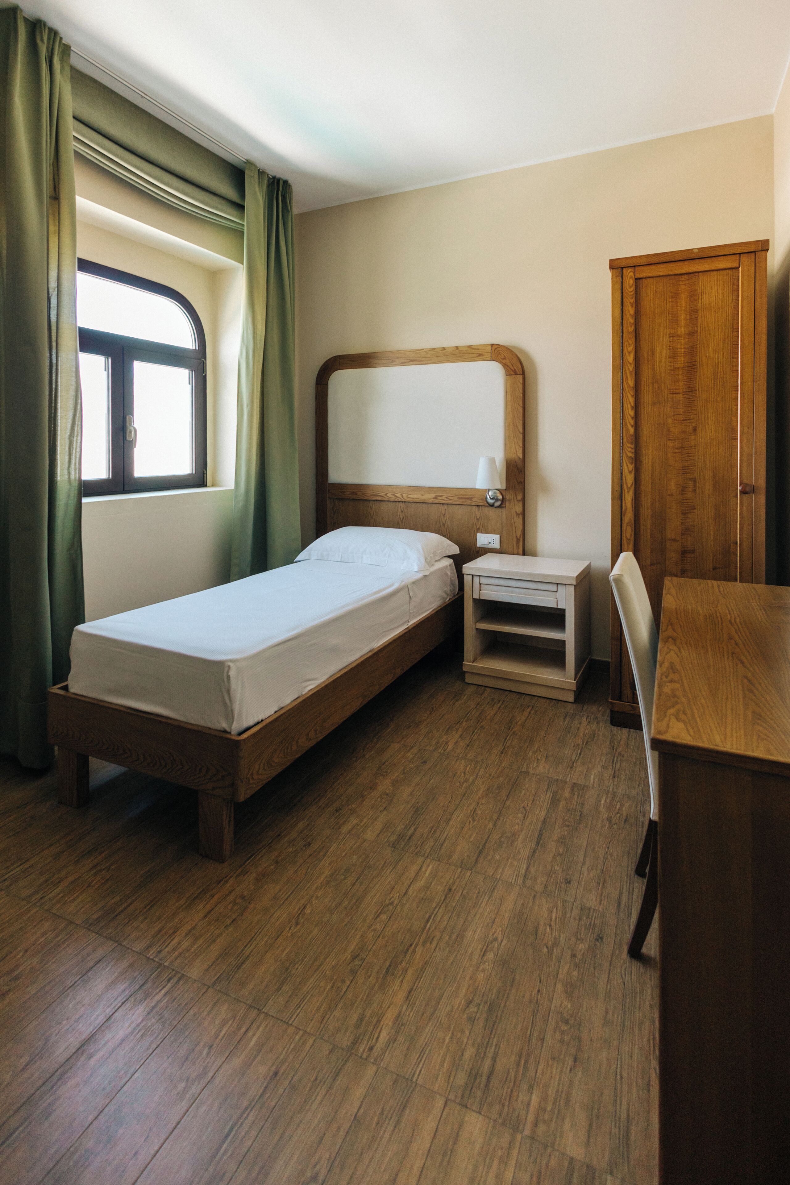 A single bed with a wooden headboard, a bedside table, a desk, and a wardrobe in a room with wooden flooring and a window with curtains.