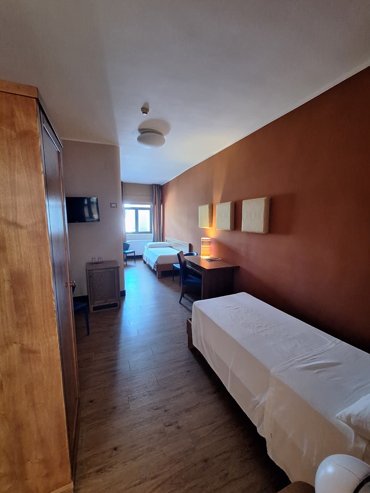 A hotel room with a wooden wardrobe, a desk with a lamp, a bed, and a television on the wall.