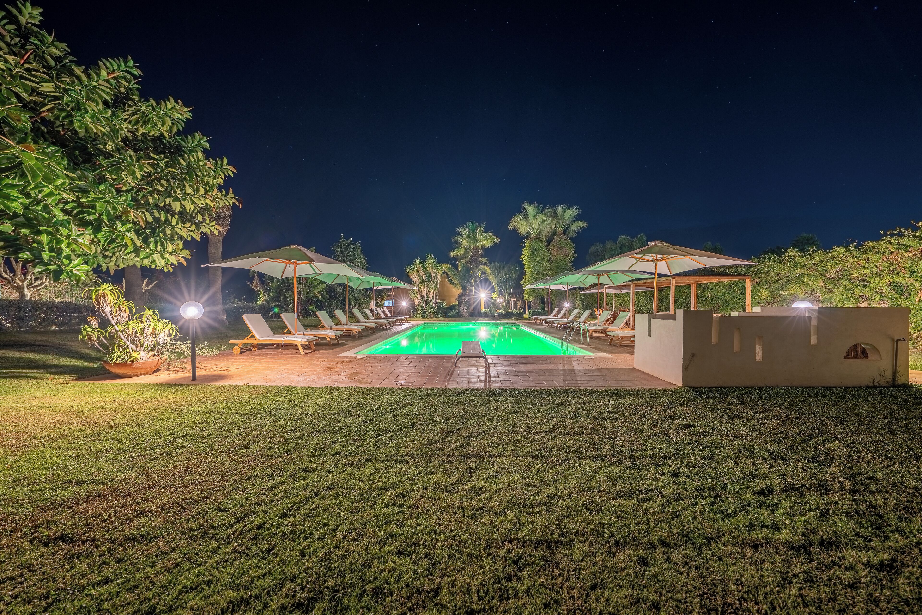 A nighttime pool area with lounge chairs and umbrellas, surrounded by greenery.
