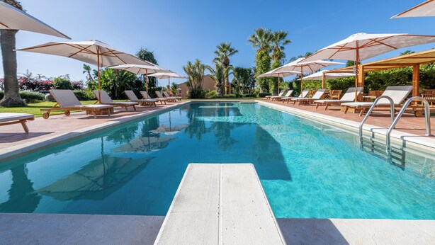 A swimming pool with lounge chairs and umbrellas, surrounded by palm trees and greenery. - Villa Carlotta Resort (Agrigento)