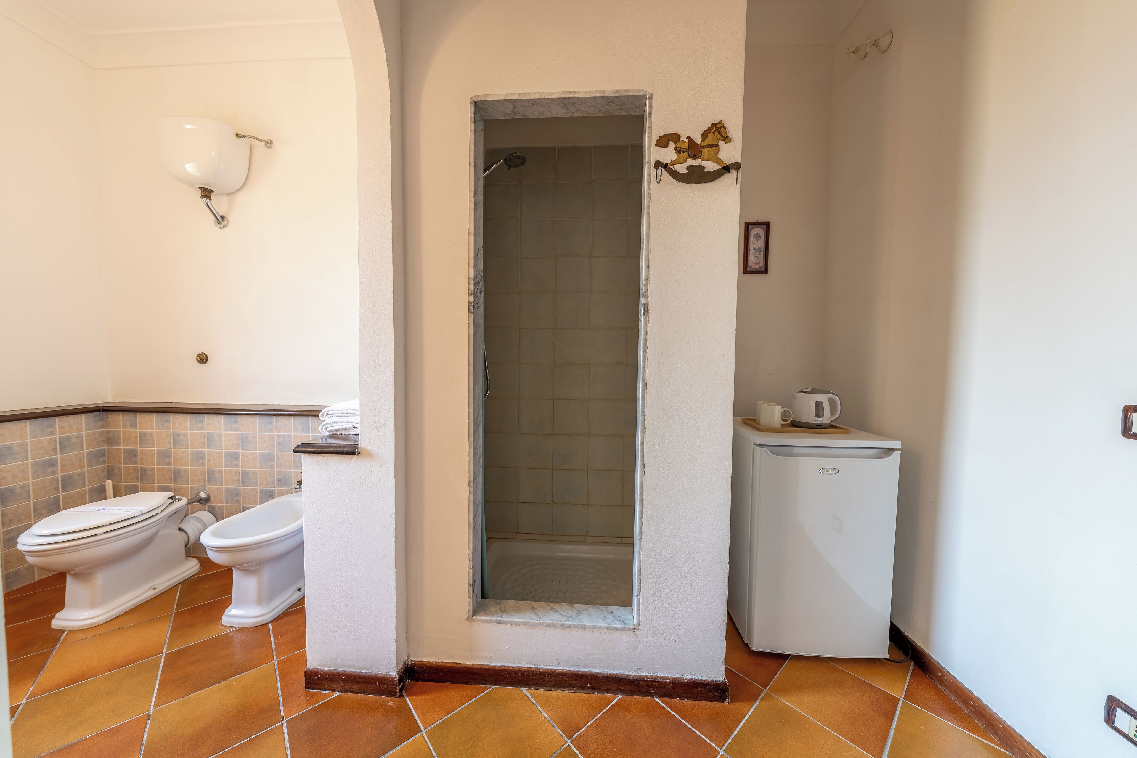 Un baño con dos inodoros, ducha y un refrigerador pequeño blanco.