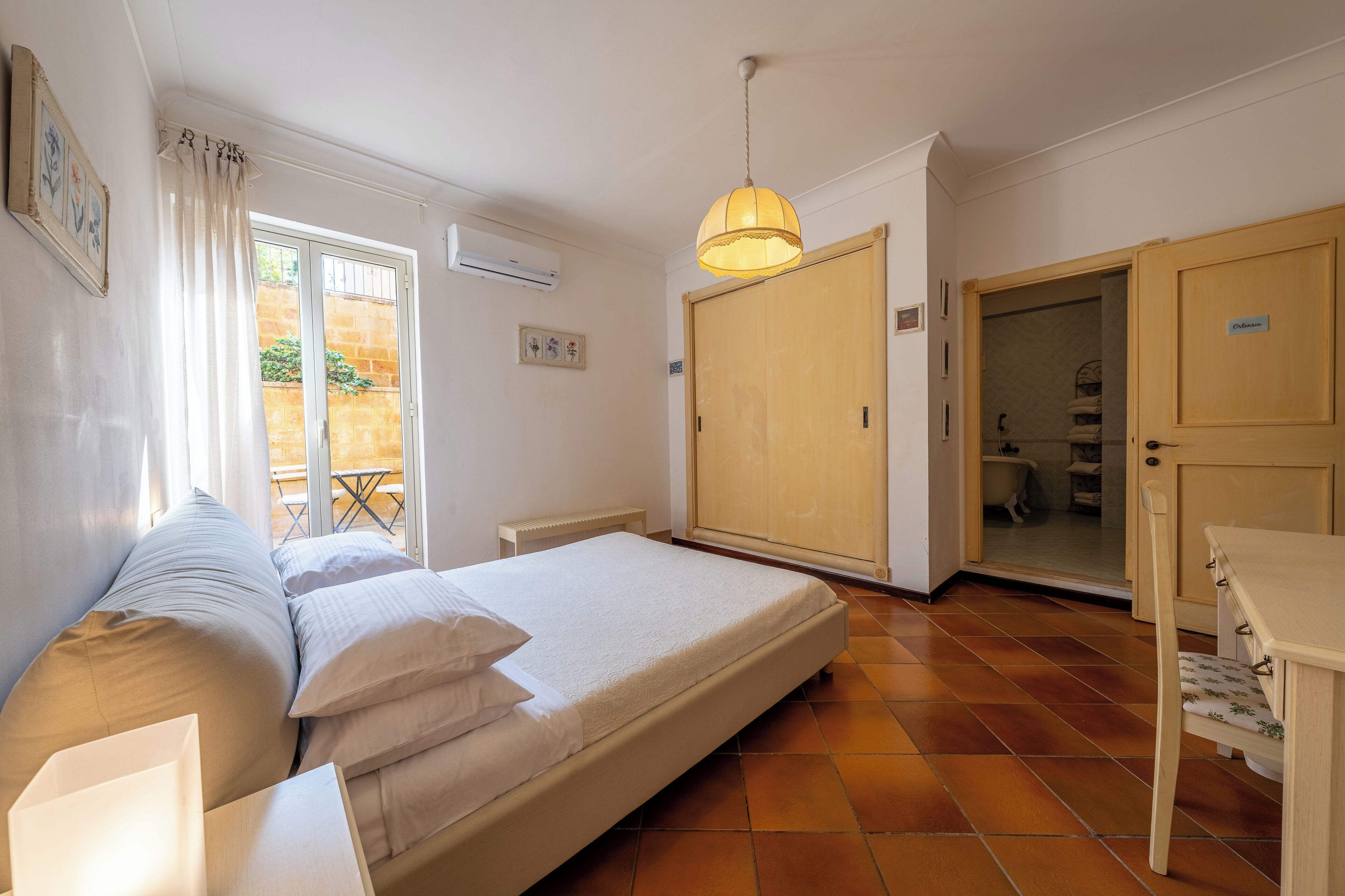 A bedroom with a bed, a wooden wardrobe, a small table, and a hanging light fixture.