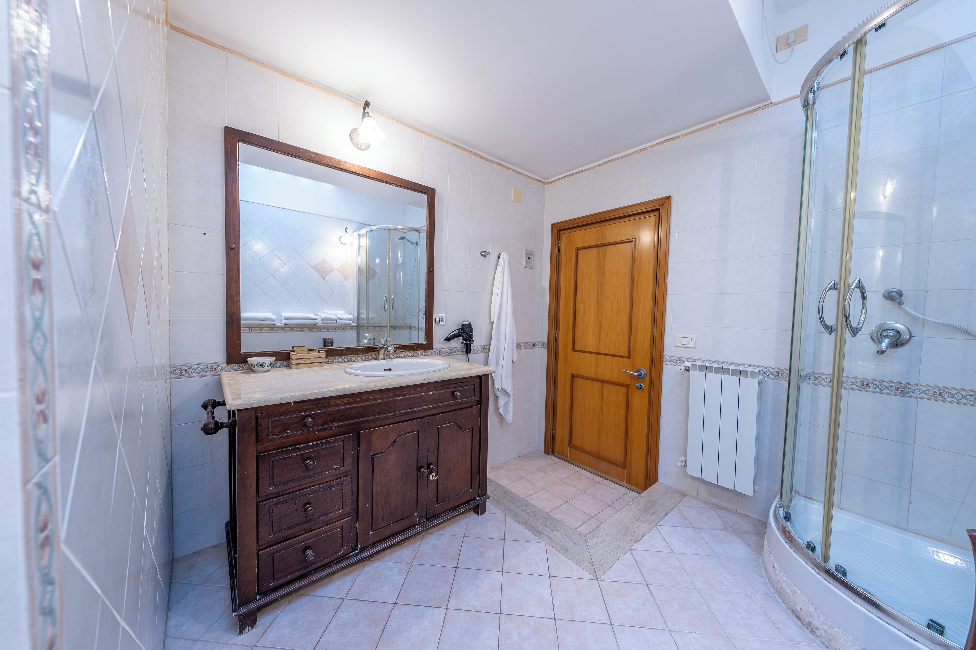 A bathroom with a wooden vanity, a large mirror, a glass shower enclosure, and a tiled floor.