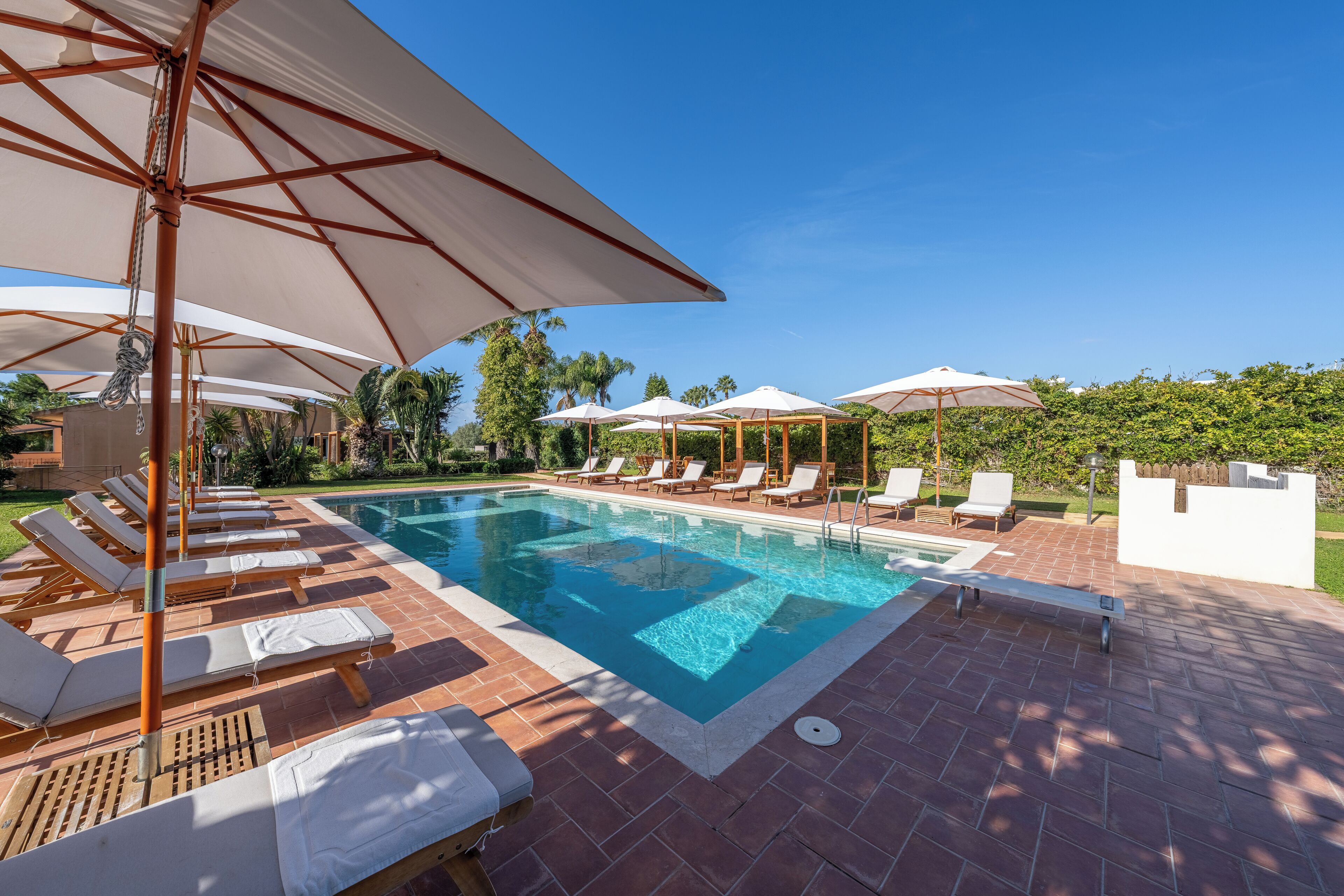 Un espace piscine avec des chaises longues et des parasols, une piscine à l’eau cristalline et une terrasse en briques.
