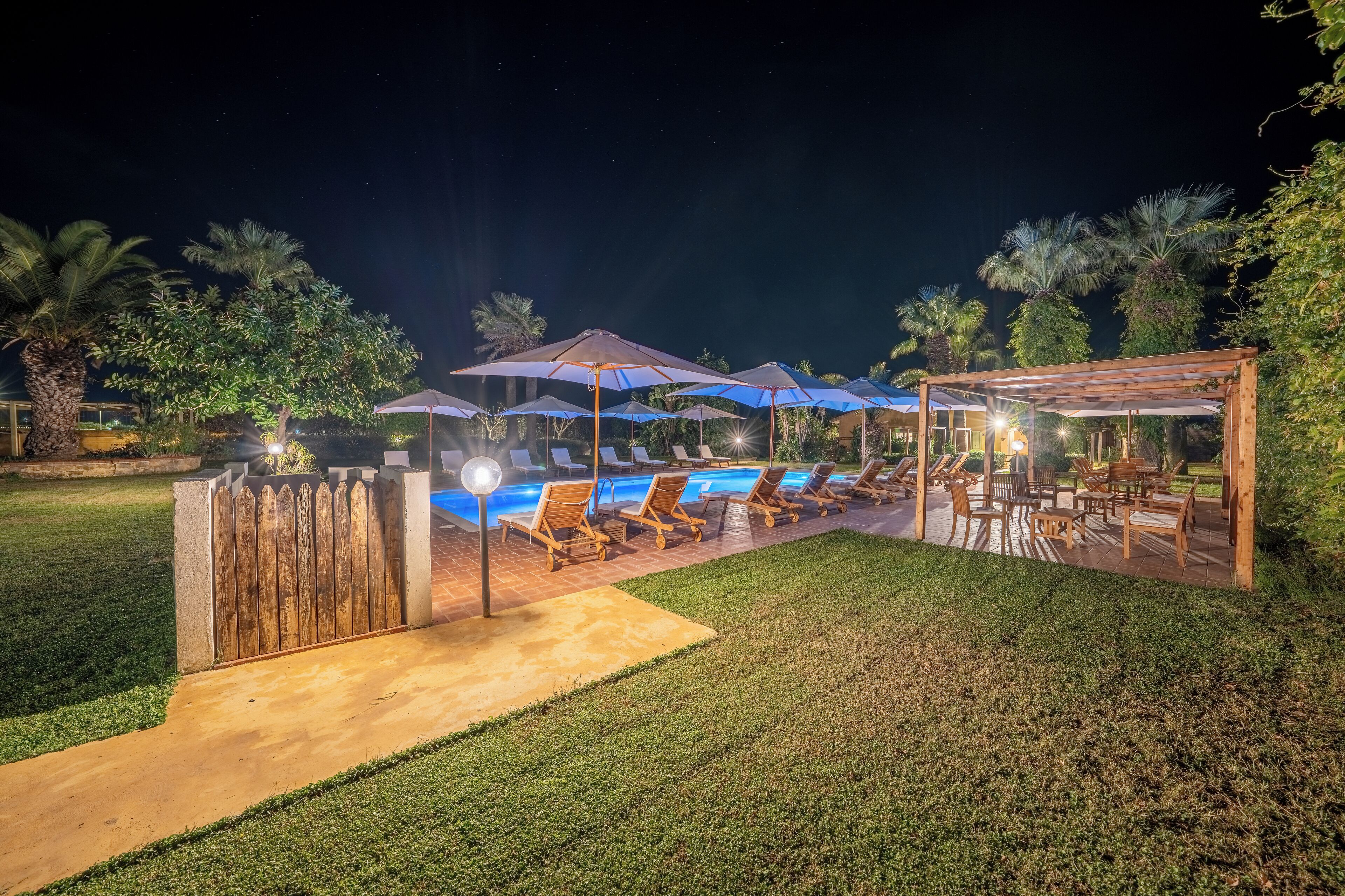 A nighttime outdoor pool area with lounge chairs and umbrellas, surrounded by a wooden pergola and a wooden fence.