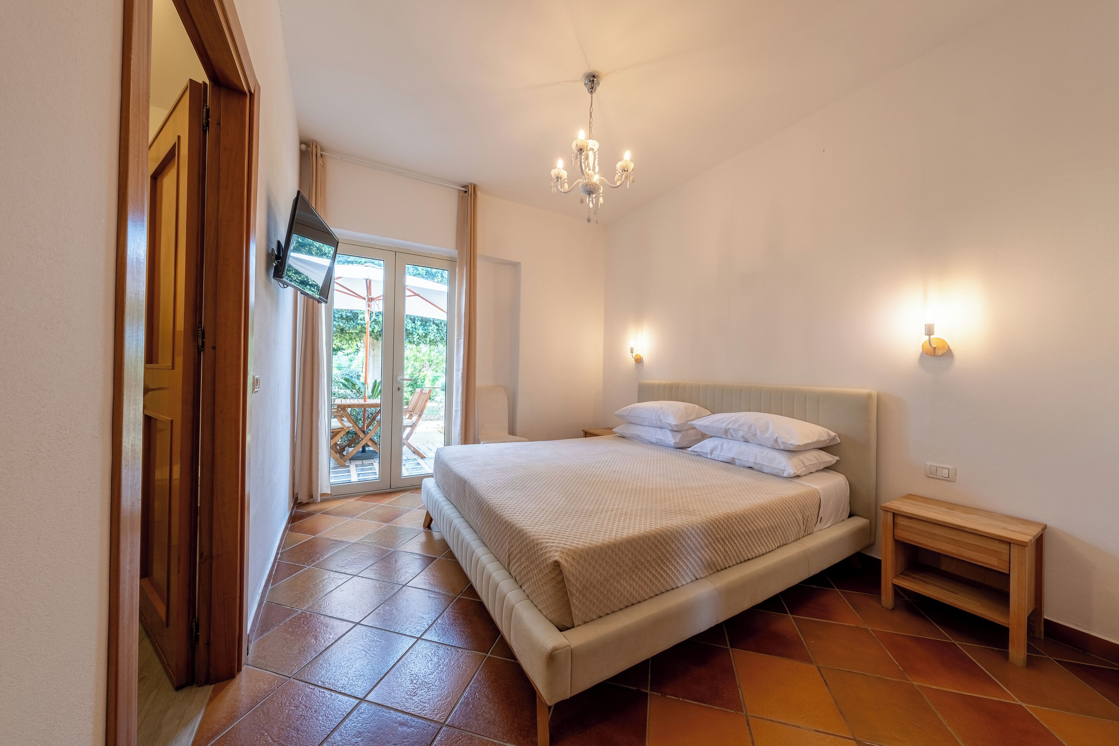 A bedroom with a bed, a bedside table, a chandelier, and a TV mounted on the wall.