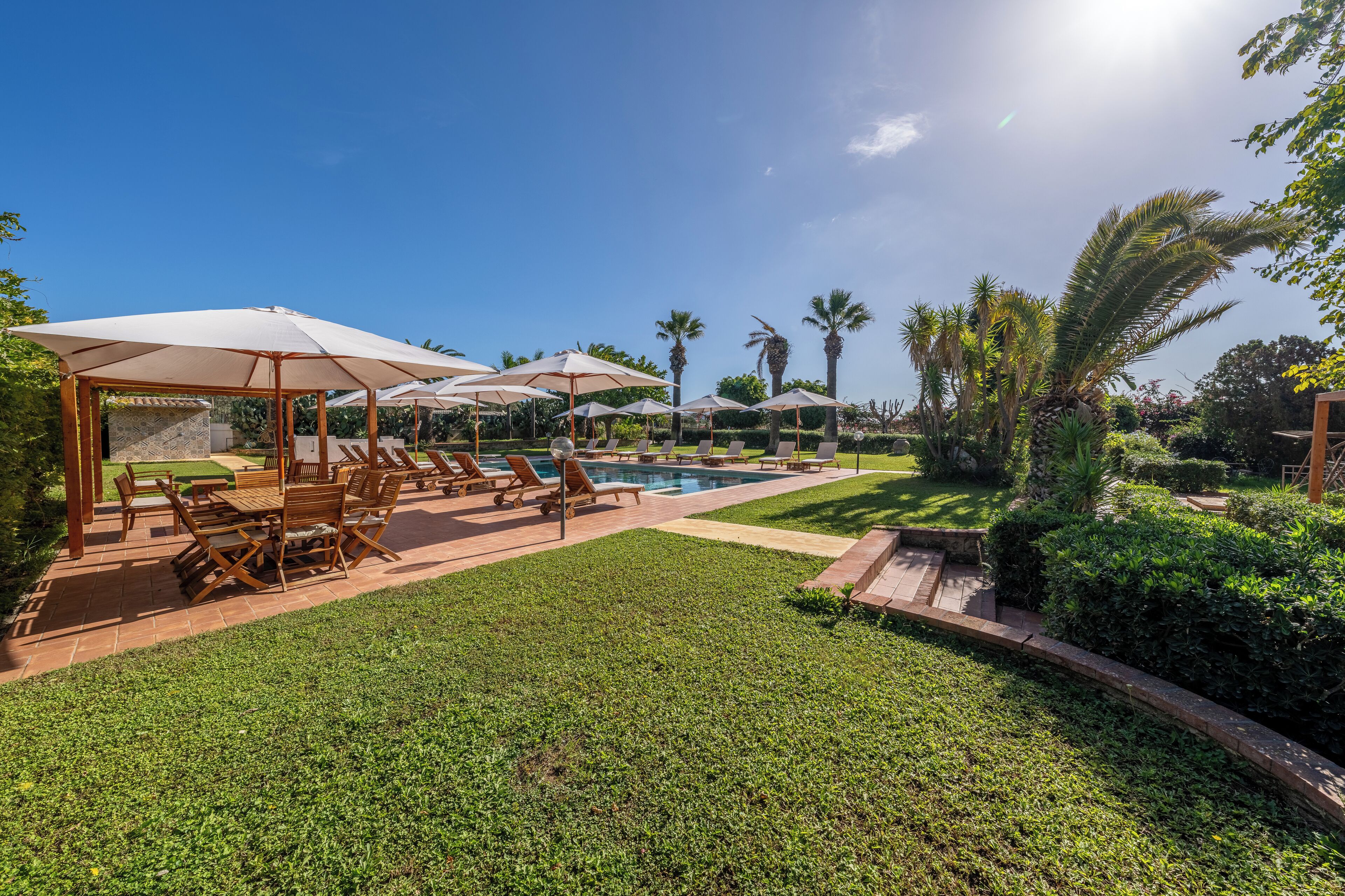 Zona de piscina con sillones y sombrillas, rodeada de vegetación exuberante y palmeras.