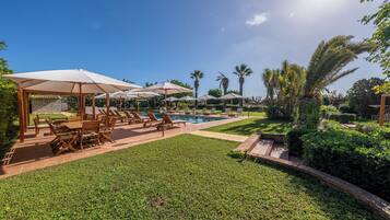 Zona de piscina con sillones y sombrillas, rodeada de vegetación exuberante y palmeras.