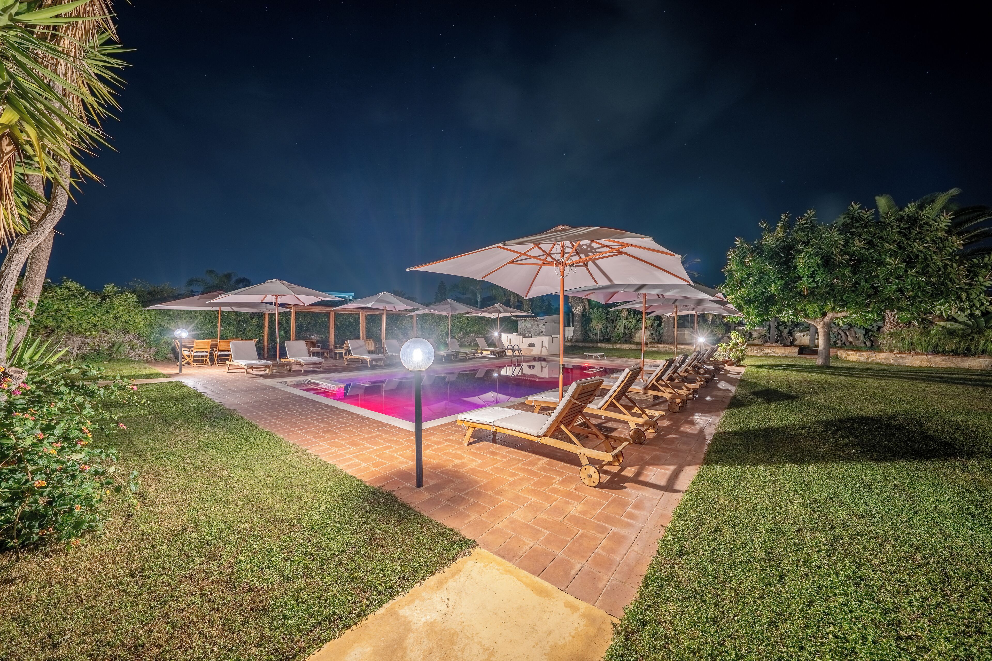 Un espace extérieur avec piscine, aménagé pour la nuit, comprenant des chaises longues et des parasols, le tout entouré de verdure.