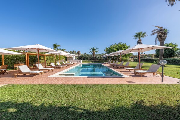 A swimming pool with lounge chairs and umbrellas, surrounded by palm trees and a well-maintained lawn. - Villa Carlotta Resort (Agrigento)