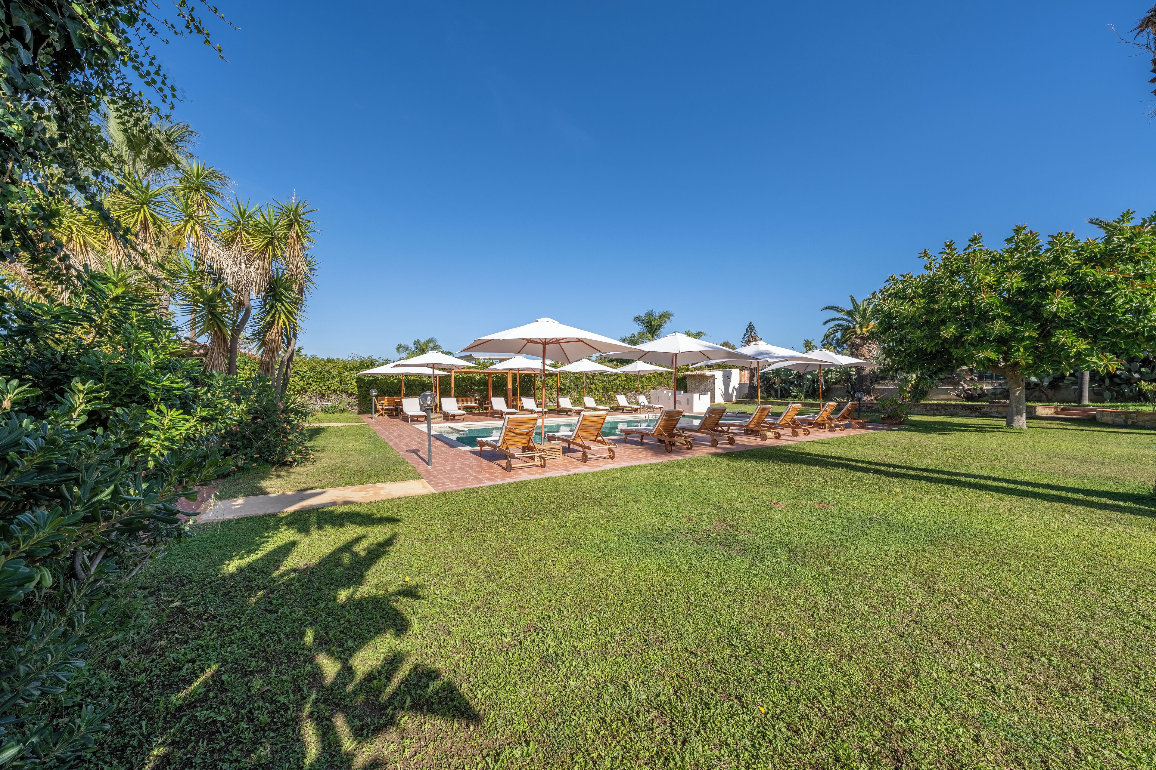 A poolside area with sun loungers and umbrellas, surrounded by lush greenery and palm trees.