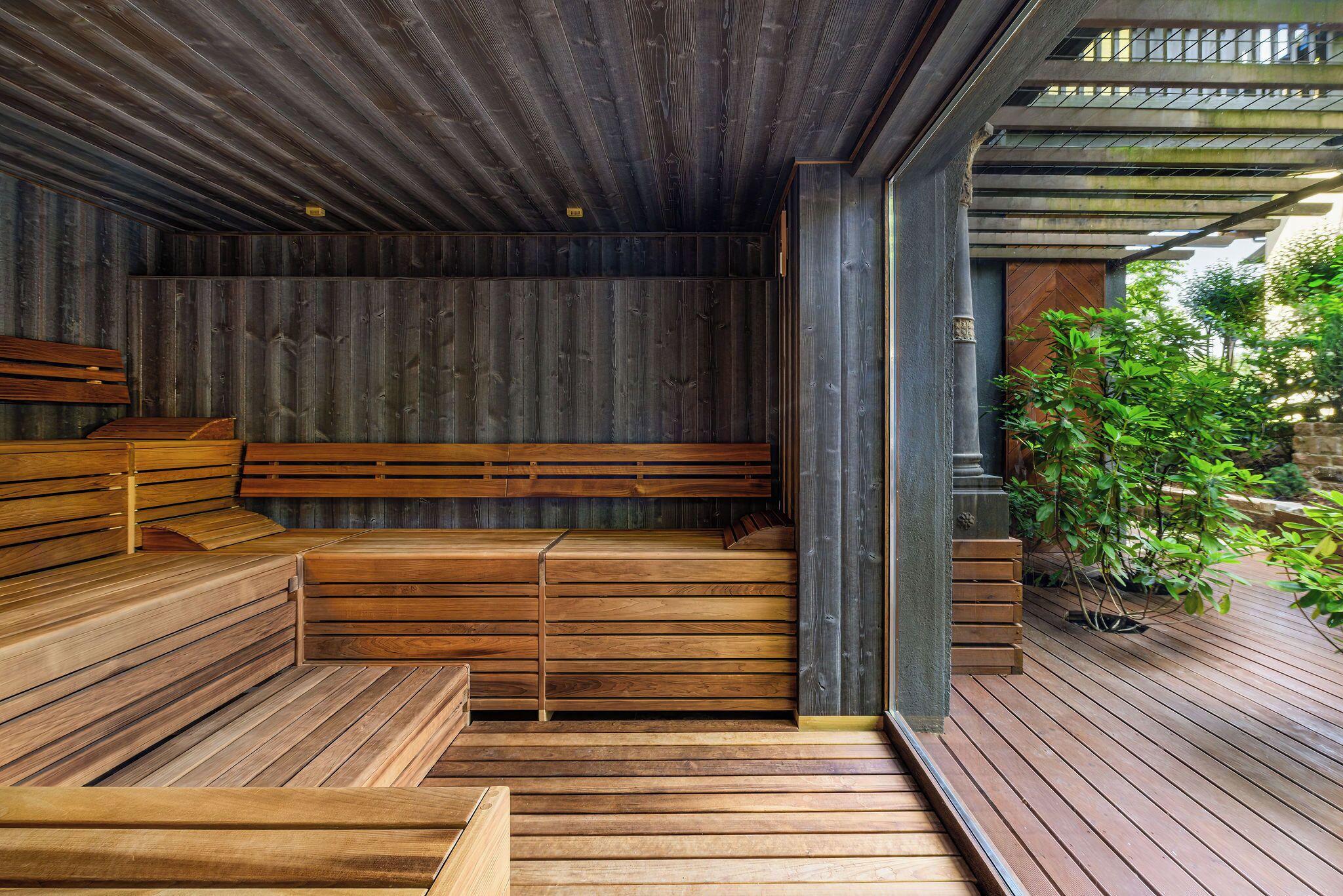 A wooden sauna with benches and a wooden floor, surrounded by a wooden deck and greenery.