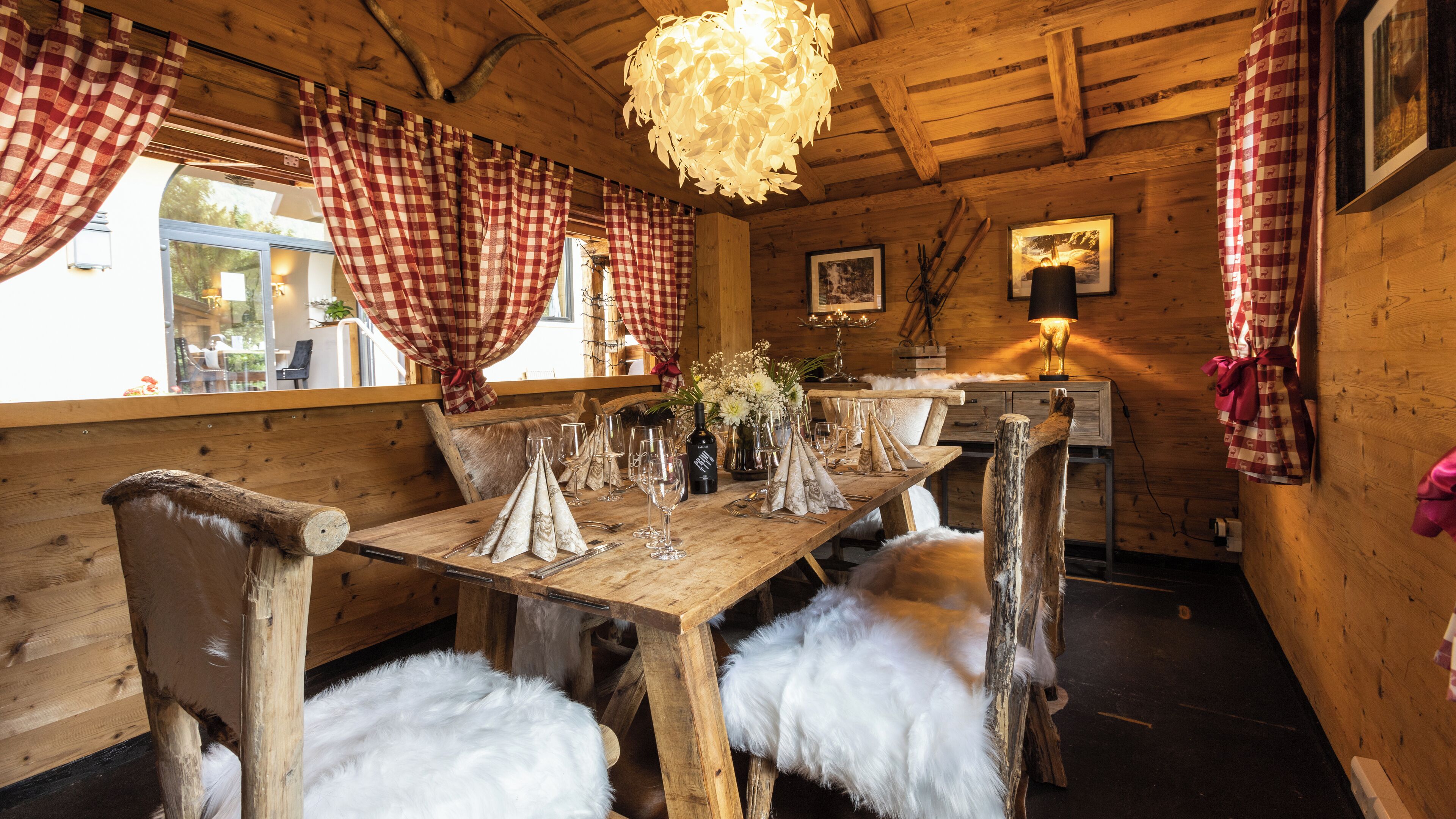 A wooden interior with a dining table set for two, fur throws on the chairs, checkered curtains, and a chandelier.