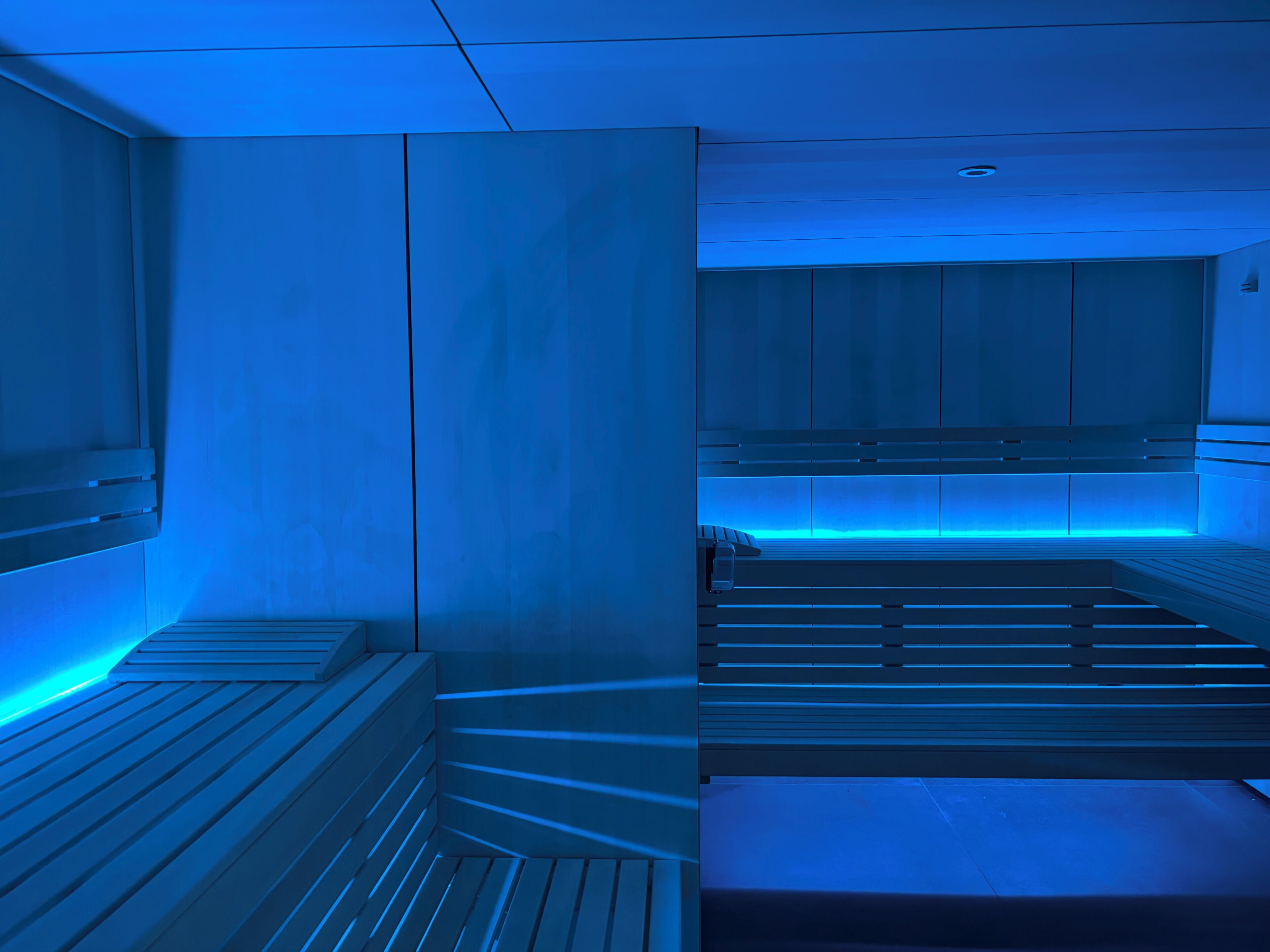 Una habitación con una camilla de masaje iluminada en azul, un banco de madera y una mesa de masaje fijada a la pared.