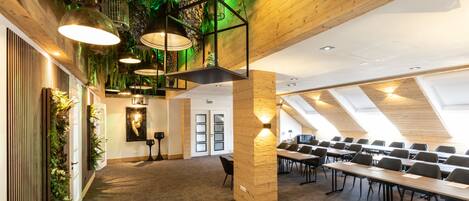 A conference room with a high ceiling, wooden beams, and a green wall with plants. There are tables and chairs arranged for meetings.