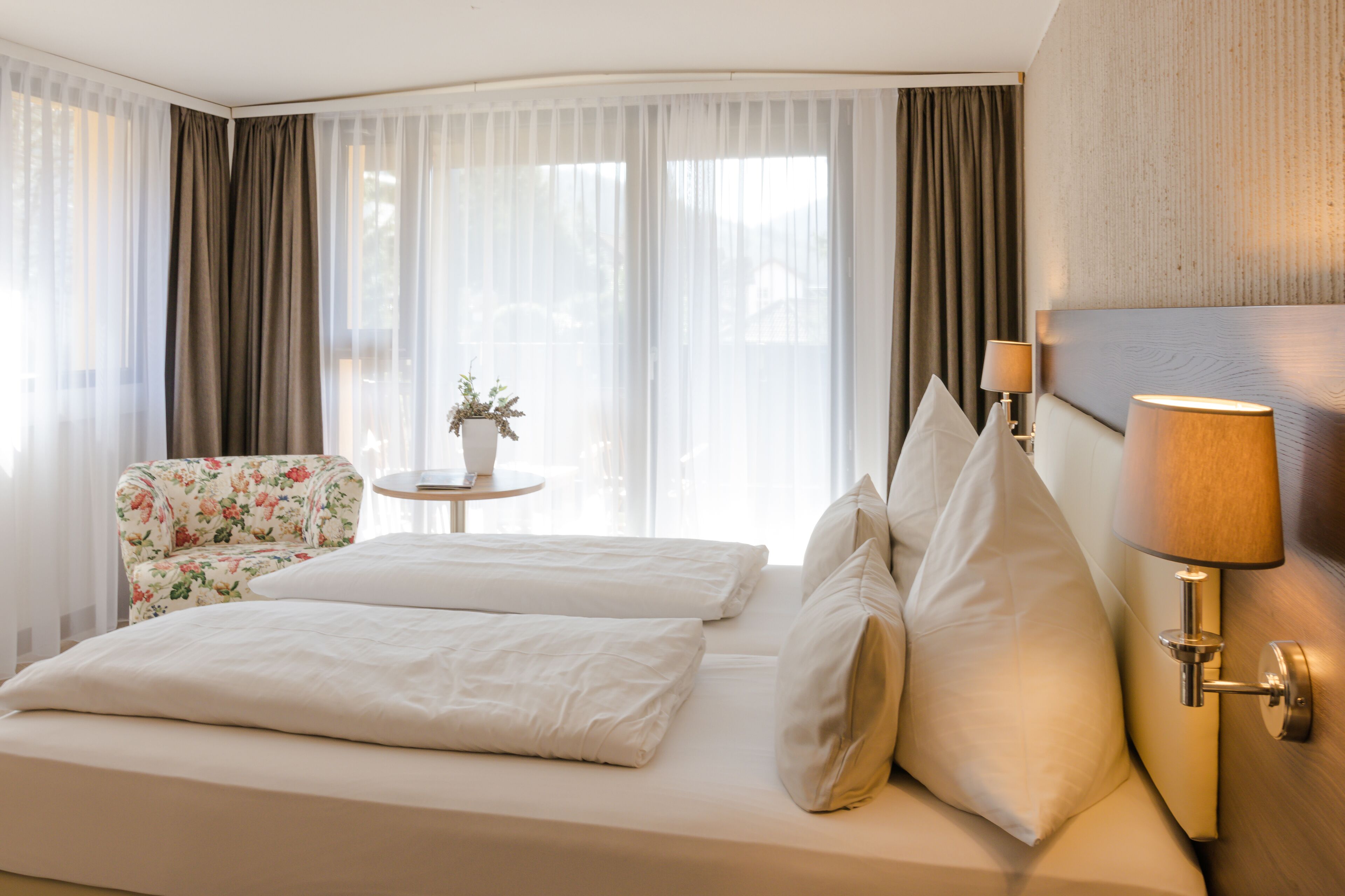 Una habitación de hotel con cama, una silla, una mesita con una planta y una lámpara.