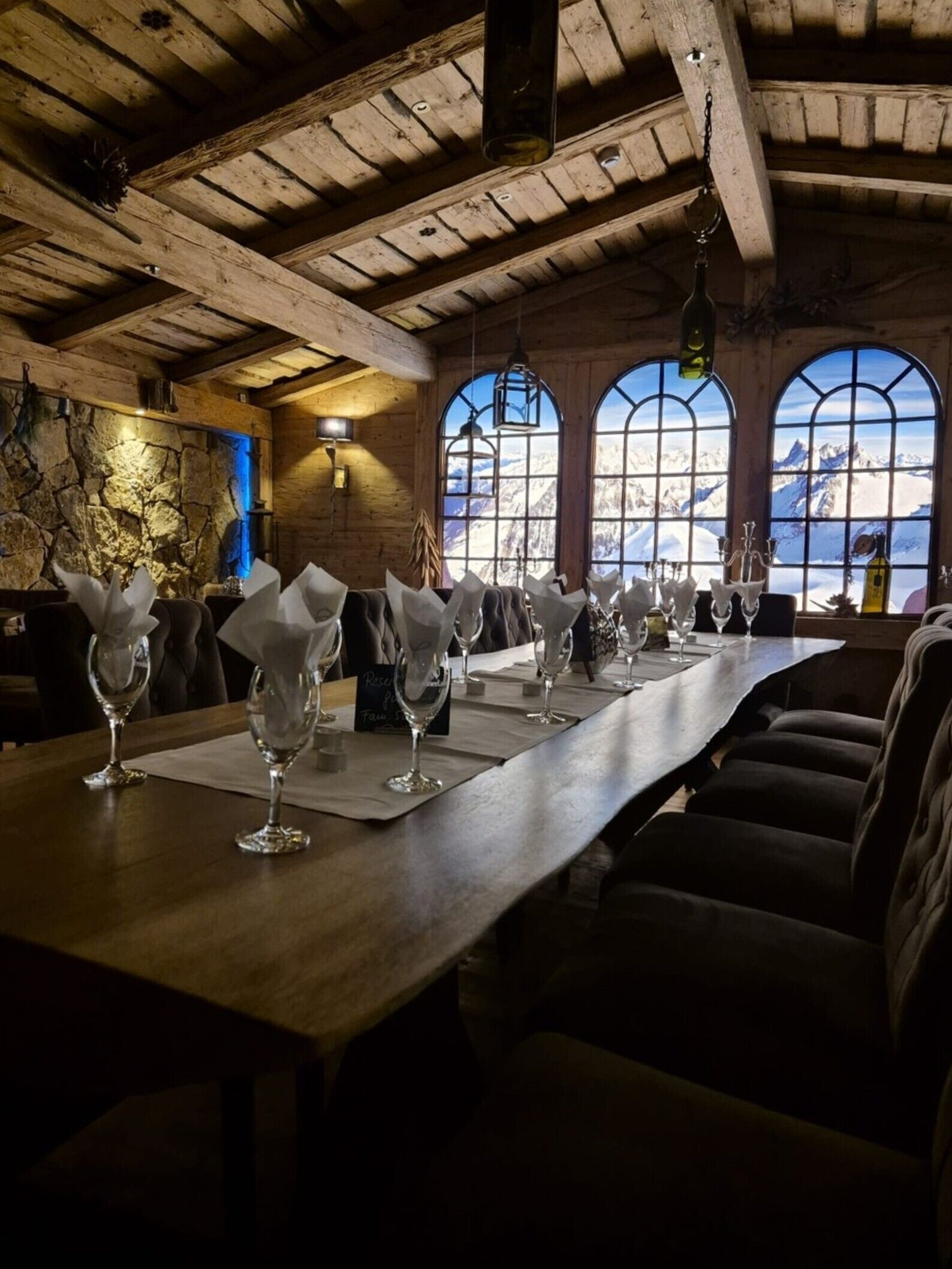 Un restaurante con una larga mesa de madera dispuesta con copas de vino, una pared de piedra y vistas a montañas a través de ventanales abovedados.